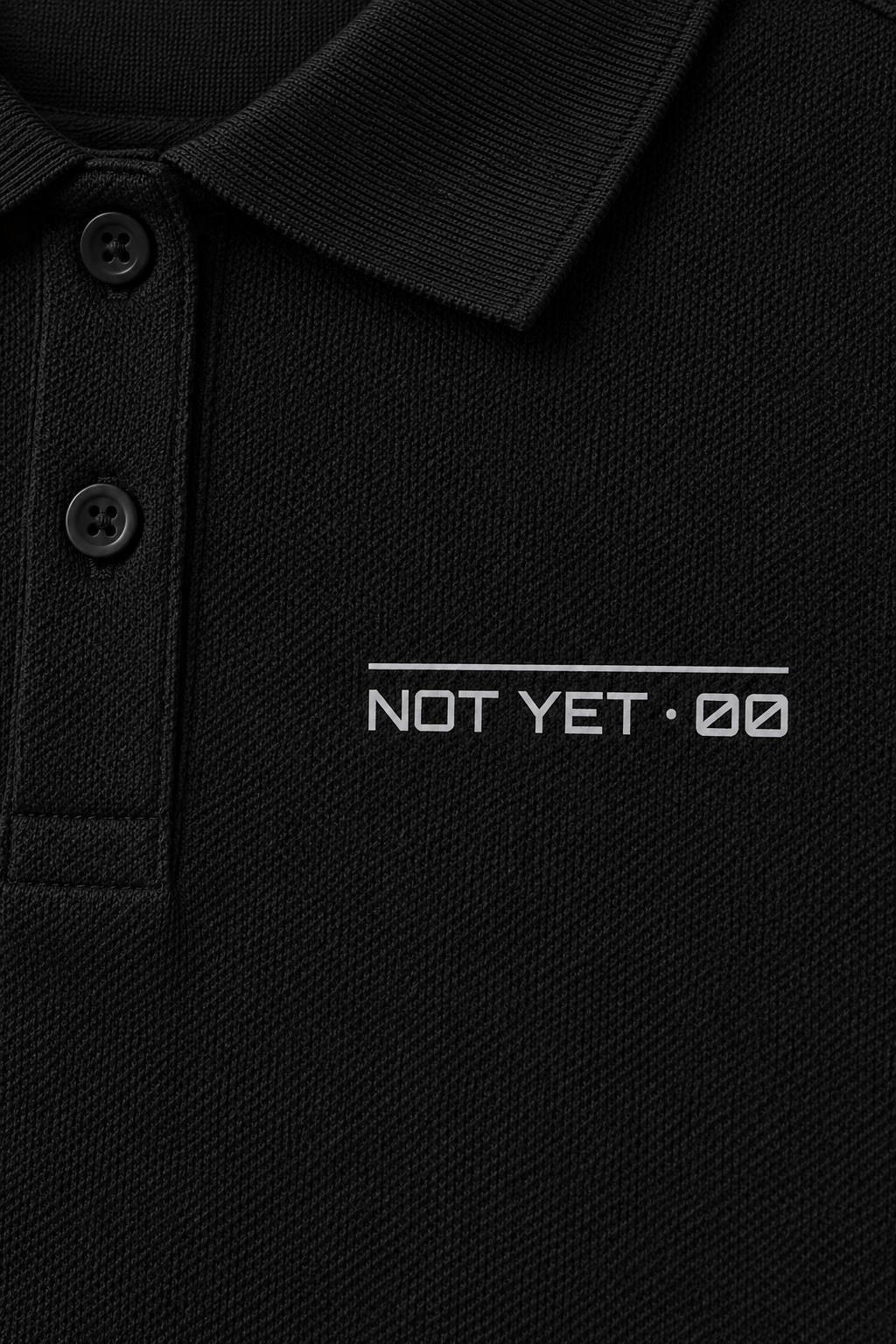 black pique polo with a small light-grey NOT YET 00 line on the chest, close-up of the collar, buttons and the small chest line