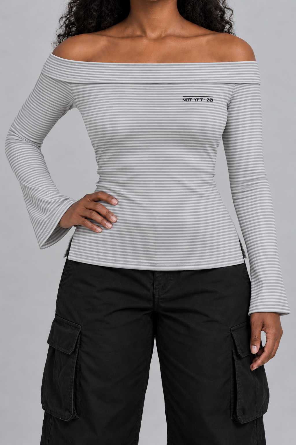 fitted off-shoulder long sleeve top in fine grey and white stripes with a tiny NOT YET 00 line on the chest, on model, front view from the collarbone down with one hand on the hip
