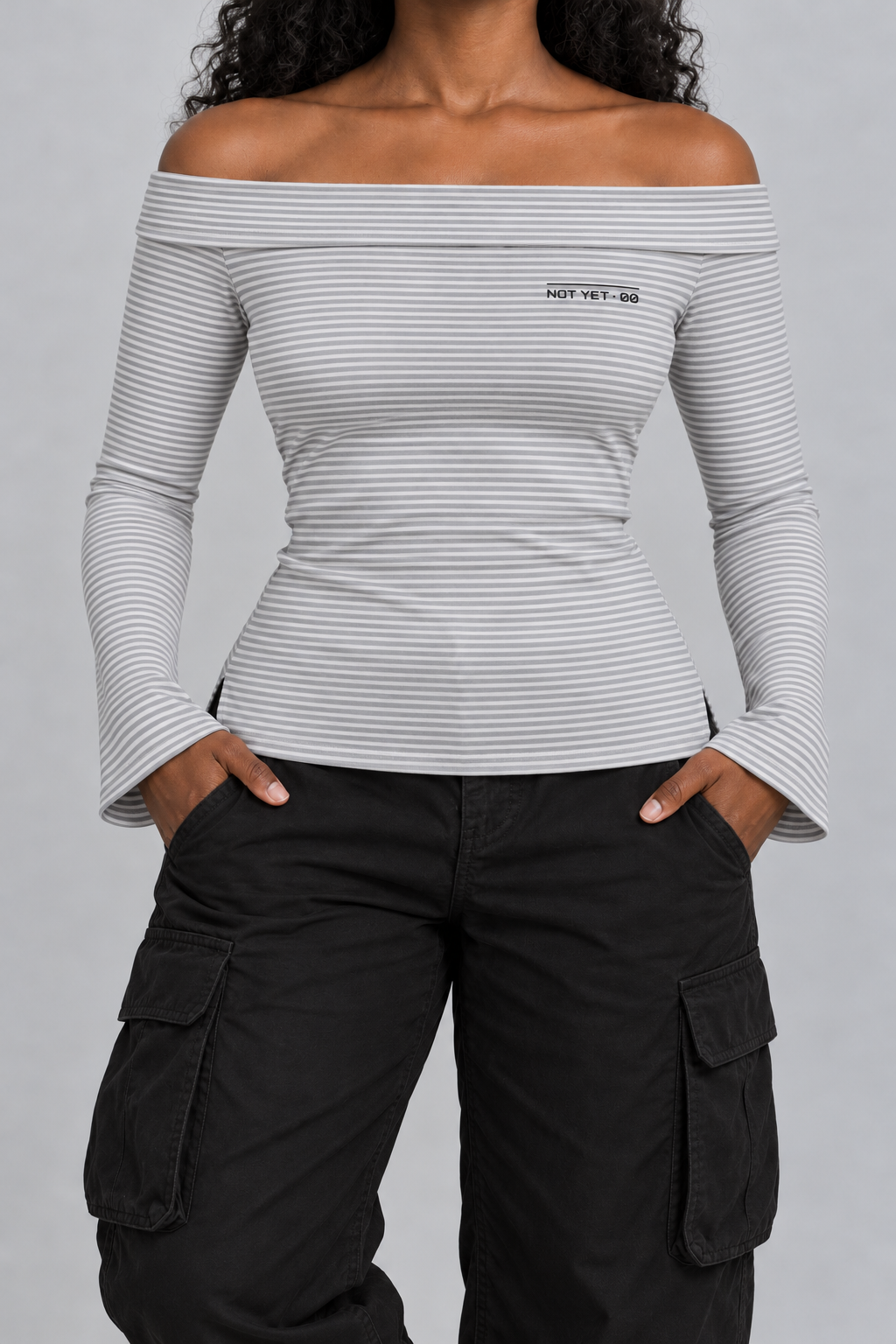 fitted off-shoulder long sleeve top in fine grey and white stripes with a tiny NOT YET 00 line on the chest, on model, front view from the collarbone down with hands in the trouser pockets