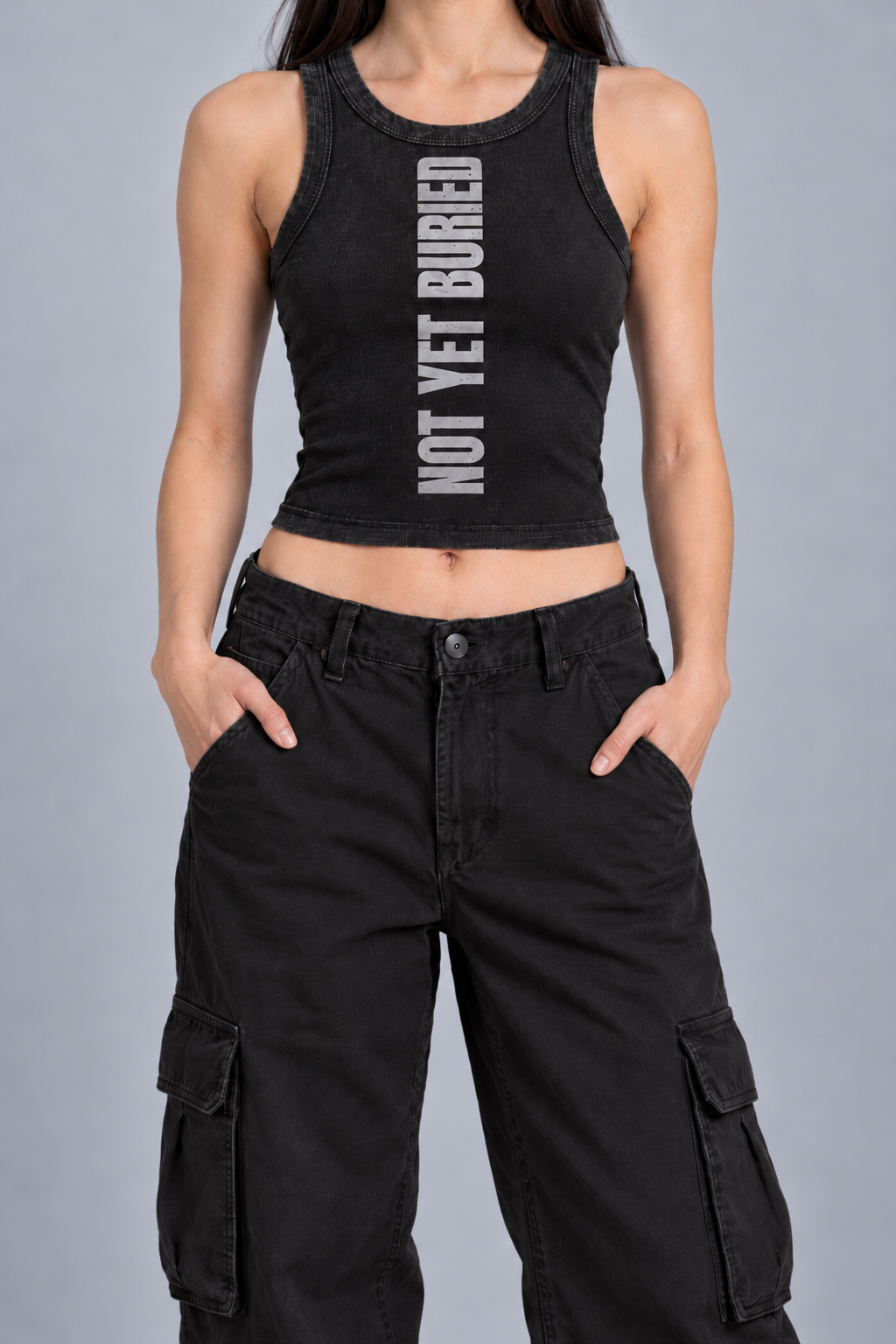 snow-washed black tank with NOT YET BURIED in tall silver-grey letters running up the front, on model, front view from the collarbone down with hands in the trouser pockets